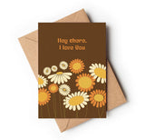A greeting card with a personal message that reads 'HEY THERE, I LOVE YOU' in combination with a floral design featuring daisies and sunflowers on a brown background, placed on top of a brown envelope.