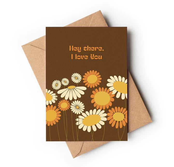 A greeting card with a personal message that reads 'HEY THERE, I LOVE YOU' in combination with a floral design featuring daisies and sunflowers on a brown background, placed on top of a brown envelope.