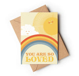 A greeting card with a colorful design, featuring a rainbow, clouds, sun, and the text 'You are so loved' on the front, placed on a brown envelope.