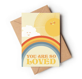 A greeting card with a colorful design, featuring a rainbow, clouds, sun, and the text 'You are so loved' on the front, placed on a brown envelope.