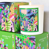 A white soy wax candle, adorned with a colorful tropical pattern, rests on a green box featuring the same design. The scene is highlighted by lush leaves and vibrant flowers in bright, assorted colors. The box bears the label "AROMA POT - LIME, BASIL & MANDARIN" along with additional script and graphic details from Aroma Pot.