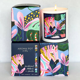 A soy candle with artwork from artist Deb McNaughton showing a colourful zebra and flowers, in a matte white vessel, surrounded by its colorful packaging.
