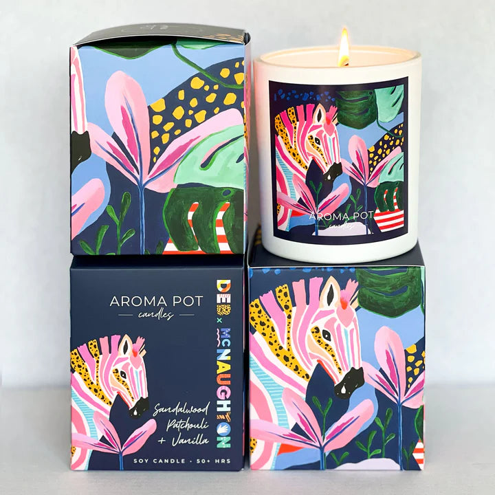 A soy candle with artwork from artist Deb McNaughton showing a colourful zebra and flowers, in a matte white vessel, surrounded by its colorful packaging.