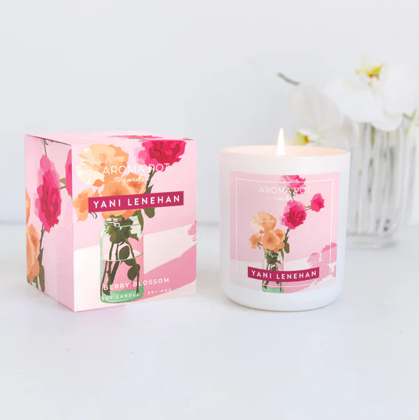 A scented candle with artwork by Yani Lenehan, pink packaging, and a lit flame, placed on a white surface with a floral arrangement in the background.