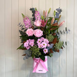 Bouquet of flowers with a pink ribbon in a clear vase against a light wooden panel background