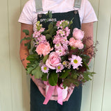 A person in a green apron holds the PETITE BLOSSOM FUSS POT by Smellies Flowers—a pink vase tied with a ribbon, filled with pastel blooms like pink roses, carnations, daisies, and greenery. Light-colored paneling is in the background.