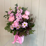 The PETITE BLOSSOM FUSS POT by Smellies Flowers features pastel pink roses, carnations, and daisies with greenery in a light pink mini ceramic vase with a ribbon, set against a pale vertical panel background.