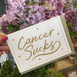 A greeting card with a beige background and gold hand-lettered text that reads 'CANCER SUCKS', surrounded by tiny hearts and a simple border. The card is held by a hand with a pink fingernail polish.