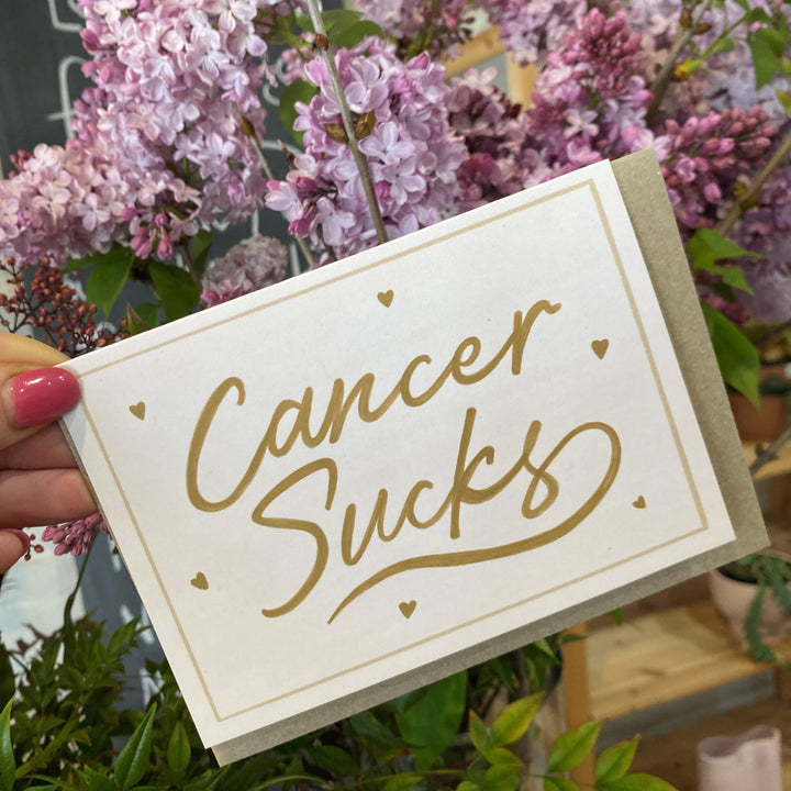 A greeting card with a beige background and gold hand-lettered text that reads 'CANCER SUCKS', surrounded by tiny hearts and a simple border. The card is held by a hand with a pink fingernail polish.
