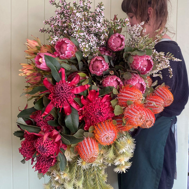A person holding a bouquet of fresh flowers including a mix of Australian and South African native flowers in a range of colors including orange, red, pink, and green.