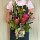 A person in a dark apron holds the FOREST FUSS POT by Smellies Flowers, a mini ceramic vase with native-inspired flowers like proteas, eucalyptus, and billy buttons, tied with a green ribbon, set against a light wooden background.