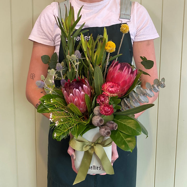 A person in a dark apron holds the FOREST FUSS POT by Smellies Flowers, a mini ceramic vase with native-inspired flowers like proteas, eucalyptus, and billy buttons, tied with a green ribbon, set against a light wooden background.