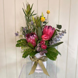 The FOREST FUSS POT by Smellies Flowers is a mini ceramic vase with “smellies,” featuring native-inspired blooms like pink proteas, yellow billy balls, eucalyptus, and greenery, tied with an olive green ribbon on light wood.