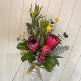 The FOREST FUSS POT by Smellies Flowers is a mini ceramic vase arrangement with pink proteas, yellow billy balls, purple thistles, pink everlasting flowers, green leaves, and eucalyptus—ideal for fans of native-inspired blooms.