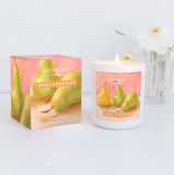 A soy wax blend candle with a cotton wick, hand-poured, adorned with a label featuring artwork of pears. The candle is displayed next to its packaging box, which also has the artwork on it, placed on a white surface with a white flower arrangement in the background.