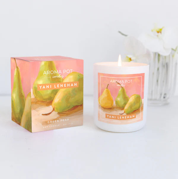 A soy wax blend candle with a cotton wick, hand-poured, adorned with a label featuring artwork of pears. The candle is displayed next to its packaging box, which also has the artwork on it, placed on a white surface with a white flower arrangement in the background.