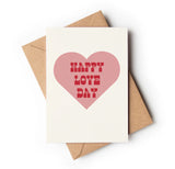A greeting card with a pink heart and 'HAPPY LOVE DAY' text in red, placed on a brown envelope.
