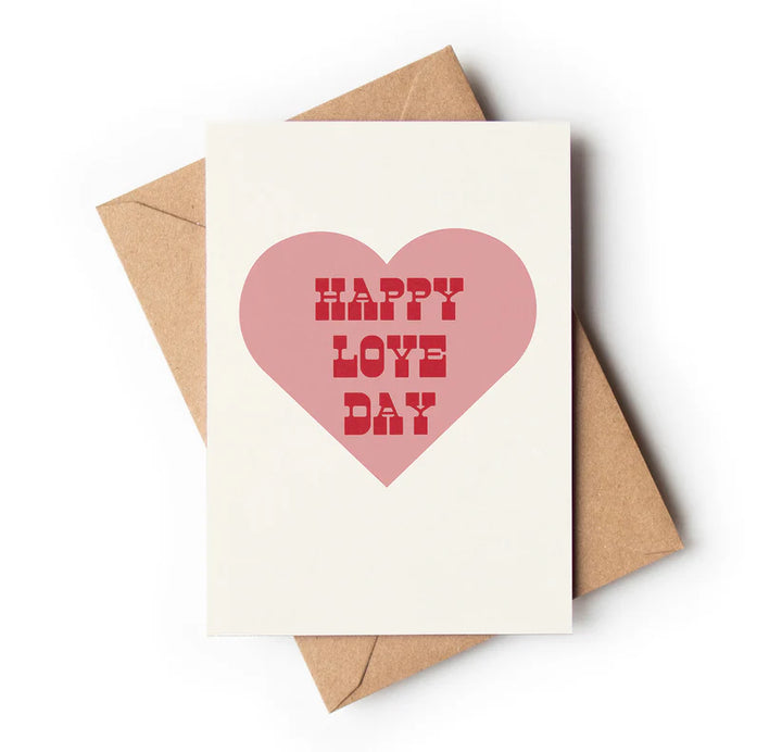 A greeting card with a pink heart and 'HAPPY LOVE DAY' text in red, placed on a brown envelope.