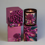 An AROMA POT - KAKADU PLUM CANDLE in a glass holder sits alongside two boxes. The candle and boxes feature a vibrant design with pink and purple flowers. The label reads "Aroma Pot" and "Kakadu Plum" against a soft gray background, hinting at the fruity fragrance within its soy wax.