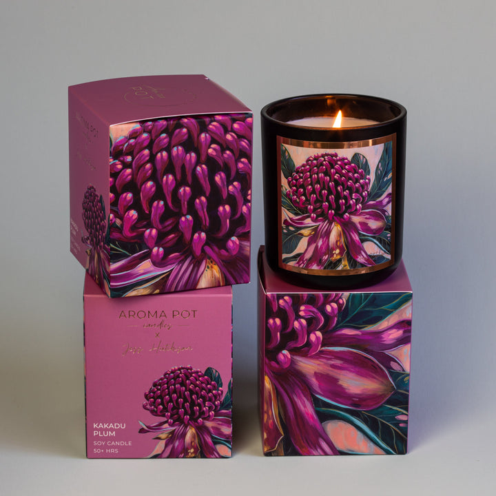 An AROMA POT - KAKADU PLUM CANDLE in a glass holder sits alongside two boxes. The candle and boxes feature a vibrant design with pink and purple flowers. The label reads