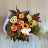 A bouquet of seasonal blooms including roses, lisianthus, and chrysanthemums with foliage, arranged in a paper wrap.
