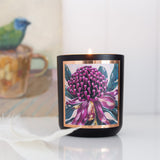 A lit AROMA POT - KAKADU PLUM CANDLE from Aroma Pot, featuring a floral illustration of a purple and pink flower on its label, is placed on a white surface. Beside it lies a feather, enhancing the serene ambiance. In the background, there is a partially blurred image of a bird perched on a cup.