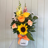 A floral arrangement featuring a selection of meadow-toned flowers including yellow, orange, and pink flowers with a sunflower, presented in a mini ceramic vase with the brand 'Smellies' visible.