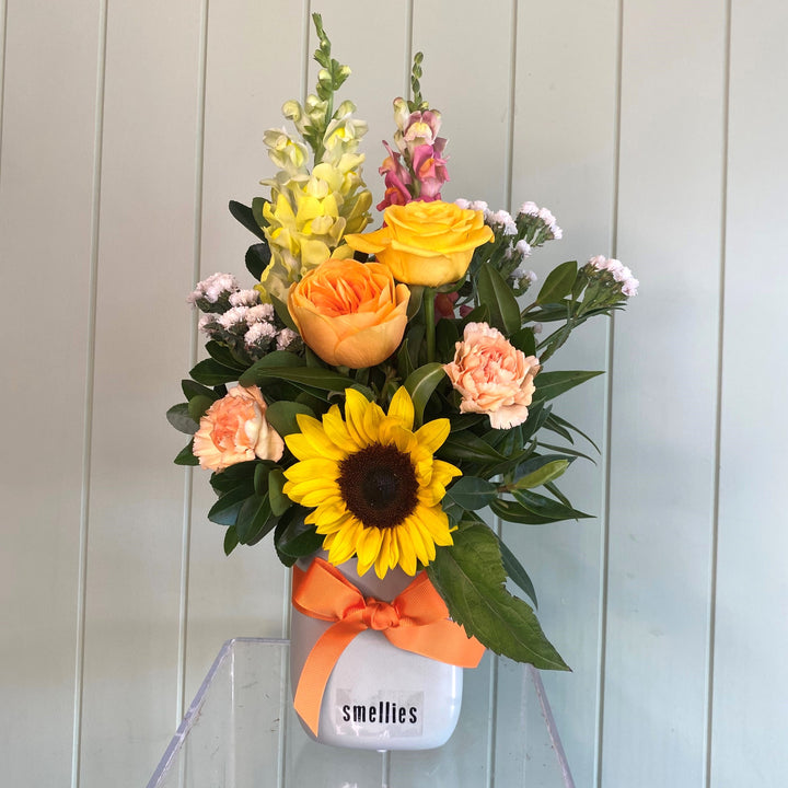 A floral arrangement featuring a selection of meadow-toned flowers including yellow, orange, and pink flowers with a sunflower, presented in a mini ceramic vase with the brand 'Smellies' visible.