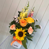 The MEADOW FUSS POT by Smellies Flowers features a vibrant sunflower, orange roses, peach carnations, yellow snapdragons, Australian blooms, and lush greenery in a vase labeled "smellies" with an orange ribbon against a white paneled wall.