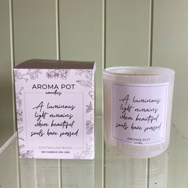 A soy wax candle with a cotton wick, scented with Australian bush, in a clear container with a label that reads 'A luminous light remains when beautiful souls have passed'. The product is displayed next to its packaging box, which has a floral pattern and the brand name 'AROMA POT'.