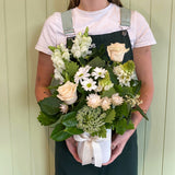 A person holding a bouquet of flowers including white and green tones with a ribbon tied around it, in front of a light-colored background.