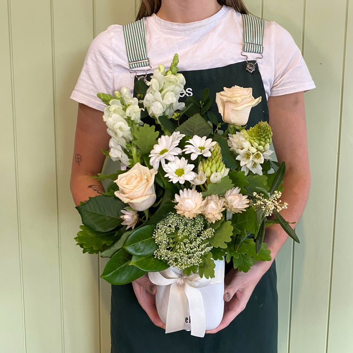 A person holding a bouquet of flowers including white and green tones with a ribbon tied around it, in front of a light-colored background.