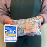 Person holding a package of Propolis Soap and a bar of soap, wearing a green apron with 'smellies' branding.