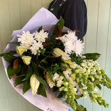 Person holding a bouquet of white flowers that includes snapdragons, fragrant lilies and chrysanthemums against a light green panelled wall.