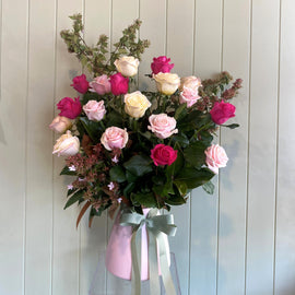 A ceramic vase filled with an arrangement of Australian grown roses in various colors including pink, white, and red, accompanied by green foliage, presented against a wooden background.