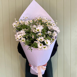 Bouquet of white daisies wrapped in paper held by a person against a light green wall.