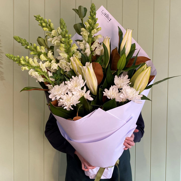 A person holding a bouquet of fragrant white lilies, chrysanthemums, and snapdragons with green leaves, wrapped in white paper.