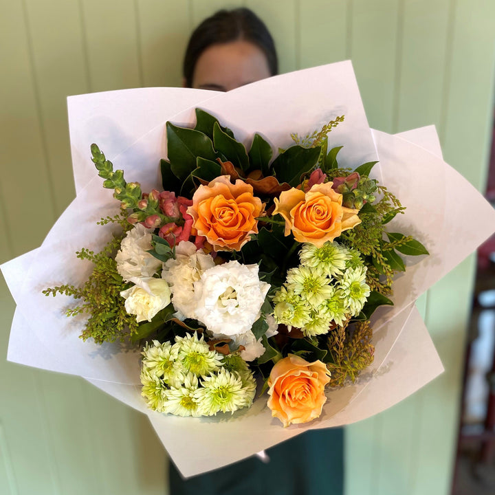 Person holding a bunch of flowers in orange, green, white and yellow standing against a wall