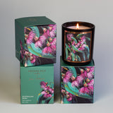 A lit Aroma Pot - Australian Bush Candle in a decorative glass holder featuring an illustration of purple flowers and green leaves. The candle is placed on its matching box, which showcases the same floral design and includes the text "Aroma Pot," "Australian Bush," and "Soy Candle 50+ Hours." Delight in the soothing scent of eucalyptus and sandalwood.