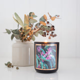 An Aroma Pot - Australian Bush Candle, crafted from soy wax and encased in a black jar with a colorful floral design, rests on a white surface. Positioned behind the Australian Bush Candle is a white ceramic vase filled with dried eucalyptus branches. The backdrop is minimalistic, featuring a light, neutral color palette.