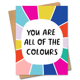 The ALL OF THE COLOURS GREETING CARD by Friends of Henry features the bold black text "YOU ARE ALL OF THE COLOURS," centered in a white oval on a vibrant, multicolored background resembling a color wheel. Bright, energetic segments splash around it, with a brown envelope subtly visible from behind.