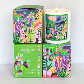 Bright and colorful soy candle next to its vibrant box. The candle label and packaging boast lively, abstract plant designs in green, pink, purple, and yellow hues. The box reads "Aroma Pot," with the scent specified as "Lime, Basil & Mandarin." This soy wax candle from Aroma Pot promises a delightful experience.