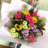 In a floral shop, a woman joyfully holds a colorful bouquet of flowers including pink roses surrounded by various floral arrangements.