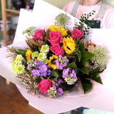 In a floral shop, a woman joyfully holds a colorful bouquet of flowers including pink roses, surrounded by various floral arrangements.