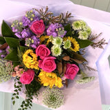 In a floral shop, a woman joyfully holds a colorful bouquet of flowers including hot pink roses surrounded by various floral arrangements.