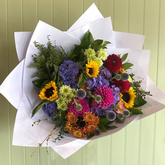 Colorful bouquet of flowers wrapped in white paper against a light green wall.