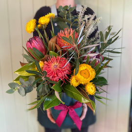 Person holding a vibrant bouquet featuring red, yellow, and orange native flowers with green foliage. The earthy bouquet is wrapped with teal and tied with a pink ribbon. The person stands against a light-colored, wooden background.