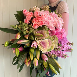 A person in a beige shirt holds a large bouquet of flowers, featuring pink roses, tulips, and ornamental cabbage, against a light green background. The seasonal arrangement includes lush green foliage and various pink and green flowers, creating a vibrant display perfect for celebrating the arrival of a baby girl.