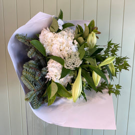 A festive Christmas-themed flower arrangement featuring white lilies, green hydrangeas, and other holiday foliage, wrapped in a white paper.
