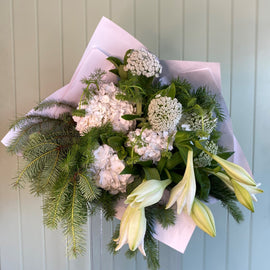 A festive Christmas-themed bouquet of flowers including lilies, hydrangeas, and other holiday foliage, wrapped in a white paper with a cardboard cone.